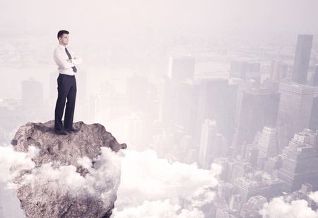 A young successful happy businessman standing confident on stone cliff in the clouds looking at the city scape from above concept