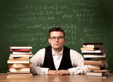 A young ambitious teacher in glasses sitting at classroom desk with pile of books in front of blackboard full of math calculations, numbers, back to school concept.