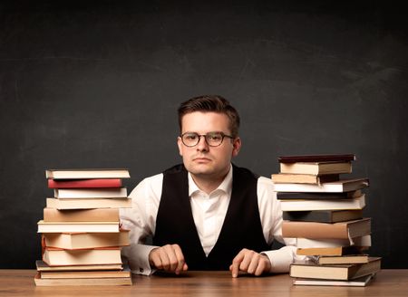 A young teacher in glasses sitting at classroom desk with pile of books in front of clean blackboard back to school concept.