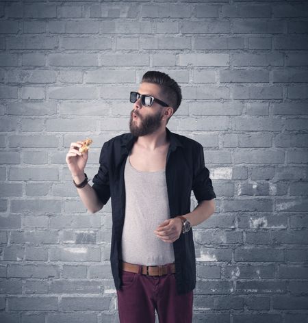 A stylish hipster guy with beard and sunglasses standing in casual clothes in front of an urban blue brick wall background concept