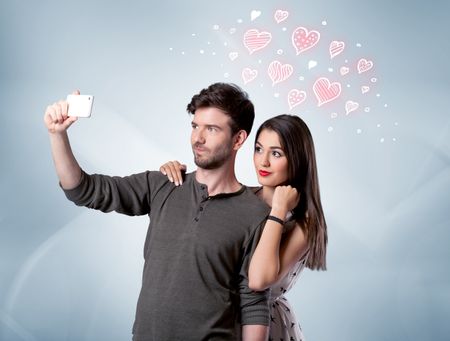A young couple in love and drawn red hearts taking selfie with a mobile  phone in the handsome guy's hand in front of an empty clear grey wall  background concept | Freestock
