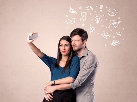 A young couple in love taking selfie with a mobile phone in the handsome guy's hand and drawn media communication icons above them, confused ideas concept