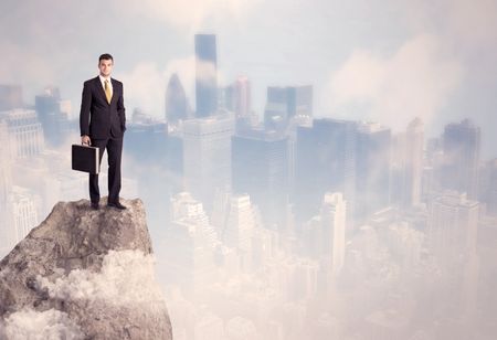 A successful good looking businessman standing on top of a high cliff above the city scape with clouds concept