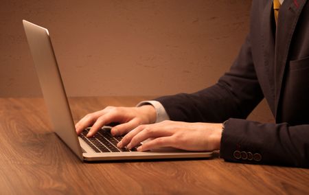 An office worker in elegant suit sitting at desk, typing on portable laptop with empty brown wall background