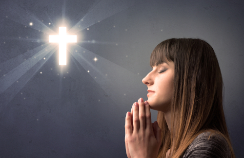 Young woman praying on a grey background with a shiny cross above her