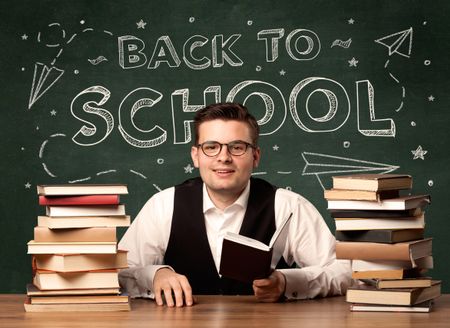 A young teacher in glasses sitting at classroom desk with pile of books in front of blackboard saying back to school drawing concept.