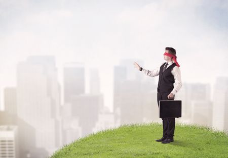 Young blindfolded businessman steps on a a patch of grass with a city in the background 