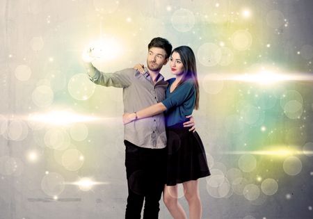 A fresh happy couple taking selfie photo with mobile phone in front of colorful lights glitter wall background concept