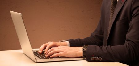 An office worker in elegant suit sitting at desk, typing on portable laptop with empty brown wall background