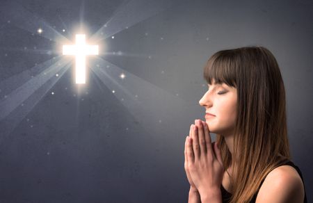 Young woman praying on a grey background with a shiny cross above her