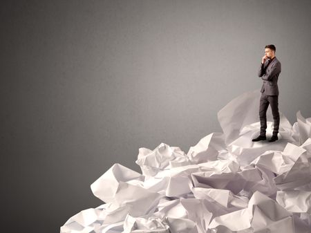 Thoughtful young businessman standing on a pile of crumpled paper with a deep grey background