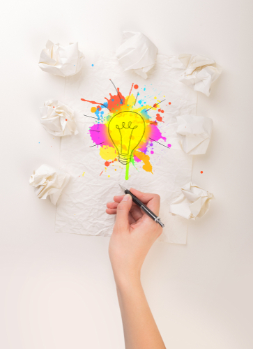 Female hand next to a few crumpled paper balls drawing a colorful lightbulb