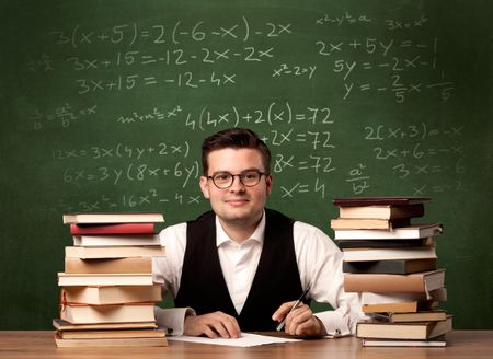 A young ambitious teacher in glasses sitting at classroom desk with pile of books in front of blackboard full of math calculations, numbers, back to school concept.