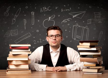 A passionate young teacher sitting at school desk with pile of books in front of blackboard drawn full of back to school items concept.