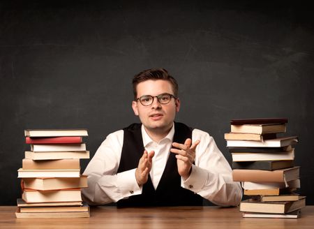 A young teacher in glasses sitting at classroom desk with pile of books in front of clean blackboard back to school concept.