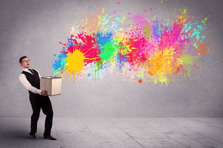 A young smiling business male holding a paperboard box with illustration of colourful spray paint splash on urban wall background concept.