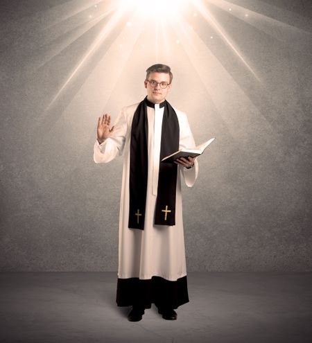 A male religious young priest in black and white dress giving his blessing, holding the holy bible while being illuminated from strong light beams coming from above concept