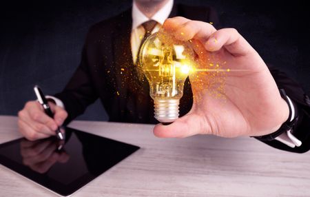 An elegant office worker holding a yellow sparkling light bulb in his hand while working in front of dark blue background concept.
