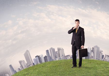 A successful male good looking business man standing in small green grass in front of city landscape with skyscrapers concept.