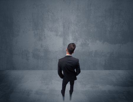 A young businessman with briefcase standing in blank empty space facing a grey urban wall, scratching his head concept.