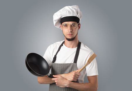 Young bearded cook portrait with kitchen tools and empty wallpaper