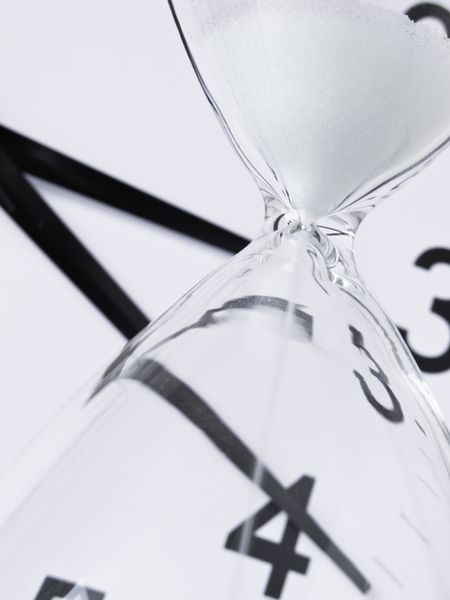 Time running out: White sand falling inside hourglass, with analog clock in background (focus on neck of hourglass), shallow depth of field