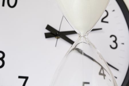 Two measurements of time: White sand falling inside hourglass, with round analog clock in  background (focus on neck of hourglass), shallow depth of field